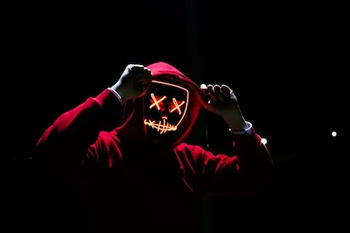 Photo of a person wearing a glowing red hooded mask with stitched features, set against a dark background.